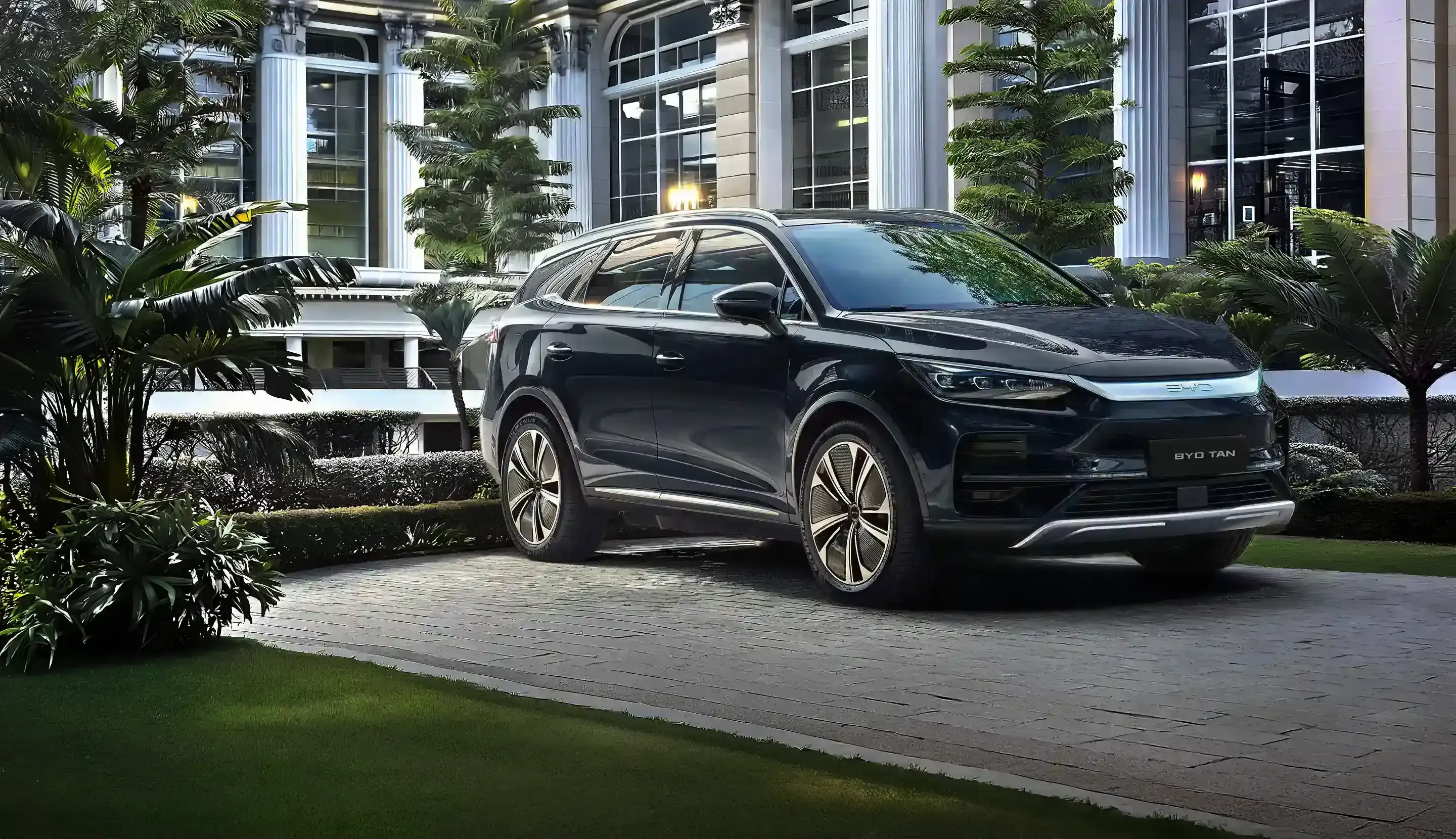 Fotografia de um SUV BYD Tan escuro estacionado em um pátio de pedras, com um elegante prédio de colunas brancas e plantas tropicais ao fundo.