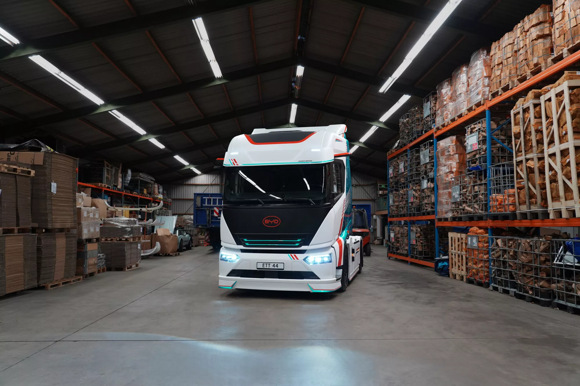 Frente do caminhão elétrico BYD ETT 44 com faróis LED ligados, estacionado dentro de um armazém de distribuição organizado, demonstrando operação de zero emissões na logística interna.