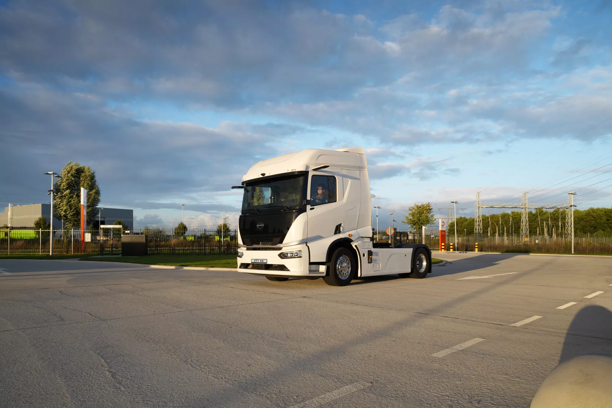 Caminhão elétrico BYD ETT 44 branco, estacionado em área gramada perto de um rio com aerogeradores e uma ponte ao fundo, mostrando a sustentabilidade no transporte
